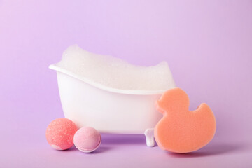 Small bathtub with foam, sponge and bath bombs on lilac background