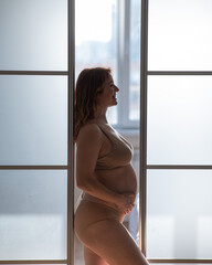A pregnant woman in nude lingerie stands in profile between folding doors.