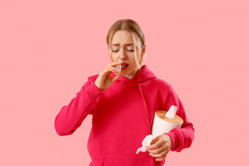 Allergic young woman with nasal drops and tissue box on pink background