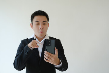 Young Asian businessman with a shocked face expression holding a smartphone by his left hand and pointing on it using his right hand