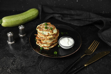 Plate of tasty zucchini fritters with sour cream on black background