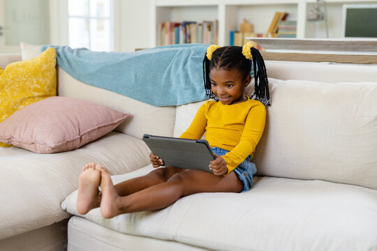 African american smiling girl using digital tablet while sitting on sofa in living room