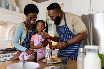 Cheerful african american man feeding fruit to daughter held by mother on kitchen counter