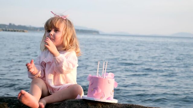 a little girl in a pink dress sits and eats a pink cake with three candles with a pen against the backdrop of a beautiful ocean sea cute grimaces squinted little, young children future birthday