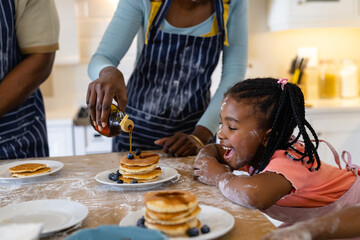 African american curious girl with mouth open looking at mother pouring honey on pancakes in plate