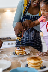 Happy african american mother and daughter pouring honey on pancakes in plate at home