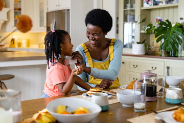 Smiling african american mother cleaning daughter's face with tissue paper at dining table