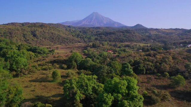 Vuelo a&eacute;reo sobre campo verde, &aacute;rboles y el Volc&aacute;n de Fuego de Colima en el horizonte. Colima, M&eacute;xico.