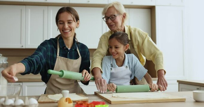 Beautiful multi generational women family wear aprons cooking together cookies, flattening prepared raw dough with rolling-pins standing in kitchen on weekend at home. Pastime, hobby, holiday recipe
