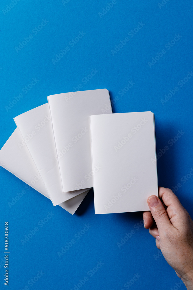 Poster Hand of caucasian woman holding a notebook over other notebooks with copy space on blue background