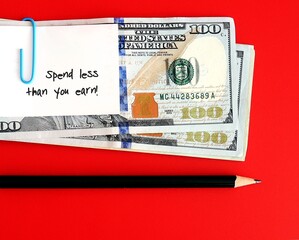 An amount of cash dollars money clipped with a white note pad written SPEND LESS THAN YOU EARN with...