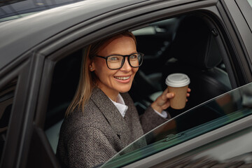 Smiling woman passenger in eyeglasses drinking takeaway coffee in taxi car on the way to work