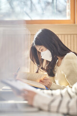 A young female Asian college student is learning or listening to a lesson while her face is outside a protective shield on the desk in a university classroom in Korea during the pandemic. This is show