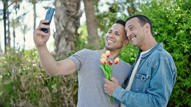 Two men couple make selfie by smartphone holding bouquet of flowers at park