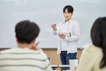 An Asian young man is standing in front of a lecture hall at a university in South Korea, giving a presentation or lecture. In front of him are male and female students. 