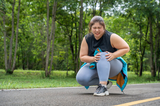 Overweight young woman with knee pain grabs her knee with both hands while sitting down at a running track of a local park