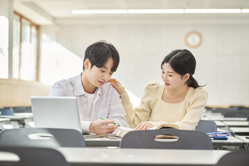 Fototapeta premium In a higher education classroom in South Korea, young university students are listening to a lecture, studying, and talking. A woman and a man are in the background