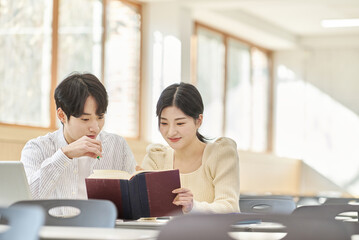 In a higher education classroom in South Korea, young university students are listening to a lecture, studying, and talking. A woman and a man are in the background
