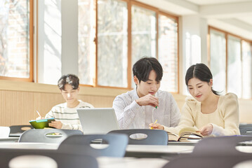 In a higher education classroom in South Korea, young university students are listening to a lecture, studying, and talking. A woman and a man are in the background