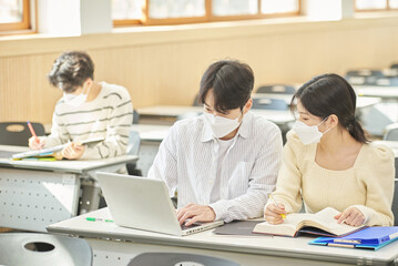 Obraz premium In a higher education classroom in South Korea, young university students wearing masks are listening to a lecture, studying, and talking. A woman and a man are in the background