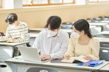 In a higher education classroom in South Korea, young university students wearing masks are listening to a lecture, studying, and talking. A woman and a man are in the background