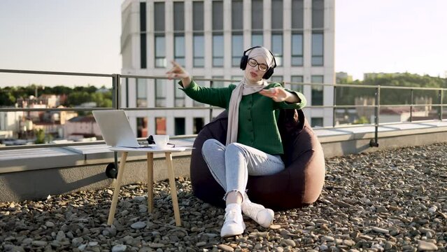Joyous arabian adult in headscarf dancing in beanbag while hearing loud tunes via hands-free headphones in open air. Female music enthusiast enjoying rich sound experience on panoramic terrace.