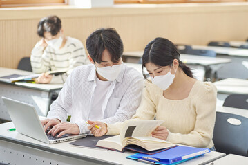 In a higher education classroom in South Korea, young university students wearing masks are listening to a lecture, studying, and talking. A woman and a man are in the background