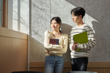 Model of a young male and female college student couple leaning against the wall of a university lecture hall in a light-filled room in South Korea, Asia.