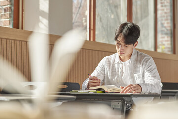 A young college student is sitting at a desk in a university classroom in South Korea, Asia. He is...