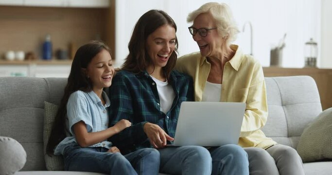 Mature granny spend time with preteen grandchild and millennial daughter laugh while watch funny on-line content, movie via streaming services sit on sofa with laptop. Amusement, family leisure, tech