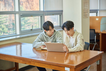 Fototapeta premium A male student sits at a desk in a university library in South Korea, Asia, looking at a laptop computer with bookshelves of books in the background.