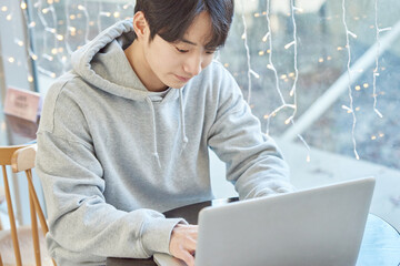 Young male student model sitting at a cafe table in Asian Korea, listening to a lecture, doing homework or working on a laptop with blurry lights in the background