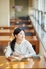 Asian Korean University Library Young female college student model sitting at a desk by a window with light coming in, reading a book, looking out the window