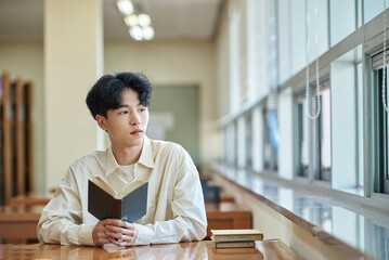 Asian Korean University Library Sitting at a desk by a window with light coming in, reading a book, looking out the window, looking toward the camera Young male college student model.