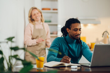 A happy interracial smart casual entrepreneur is sitting at the dining table, working on a laptop and writing in an agenda while his wife standing in the kitchen.