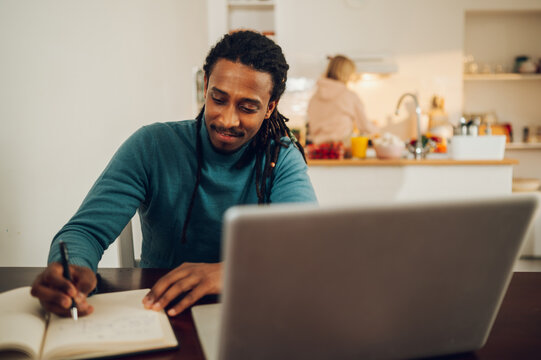 Portrait of a happy multiracial man following an online course and writing down in a textbook while his wife finishing a meal in a kitchen.