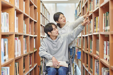 A young male college student model with a physical disability in a wheelchair looking for a book on a shelf in a university library in South Korea. A female college student model helping him