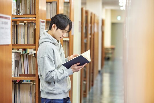 A young man attending college is browsing and reading books on the shelves with different expressions in his university library in South Korea, Asia. - Powered by Adobe