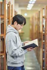 A young man attending college is browsing and reading books on the shelves with different expressions in his university library in South Korea, Asia.