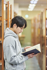 A young man attending college is browsing and reading books on the shelves with different...