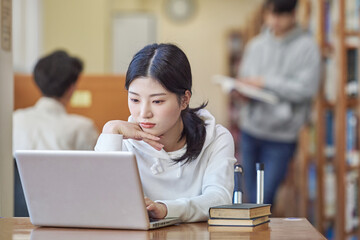 Young Asian Korean female model in library looking at laptop or book, lecture, assignment,...