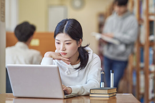 Young Asian Korean female model in library looking at laptop or book, lecture, assignment, discussion, male model in background, bookshelf in background