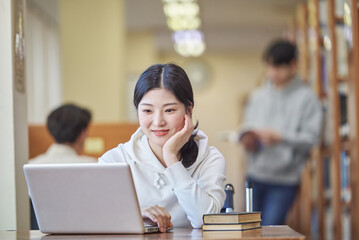 Young Asian Korean female model in library looking at laptop or book, lecture, assignment, discussion, male model in background, bookshelf in background
