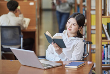 Young handicapped female college student model with disability sitting in wheelchair and reading...