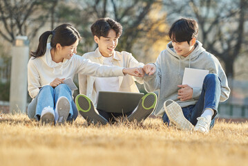 Three young college student male and female models sitting on the lawn at the university in autumn in Asian Korea, watching a laptop, listening to a lecture, discussing or talking