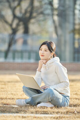 Young and attractive college student female model sitting on lawn in autumn at Asian Korean university, looking at laptop, listening to lecture, relaxing or talking online