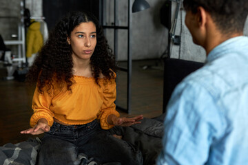 Portrait of cute african american girl of 20s with long curly hair sitting on bed in yellow top, gesticulating while telling something to her boyfriend turned back to camera, planning weekend