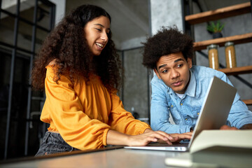 Fototapeta na wymiar Side view of charming young black happy smiling female with curly hair sitting at laptop playing video game or gambling online while her afro boyfriend looking at screen with shocked puzzled face