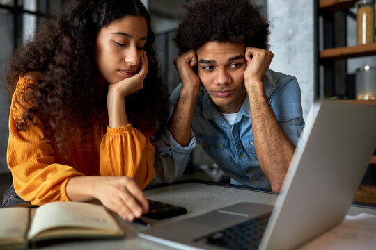 Young Married Couple Of African Ethnicity Sitting In Front Of Laptop With Puzzled Facial Expressions, Man Looking At Screen Holding Head In Hands, Female Using Calculator, Counting Expenses