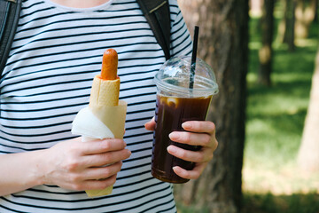 Hot dog and black iced coffee in the hands of a woman. Lunch during a summer walk in the city park.